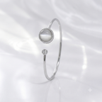 A silver-toned open bangle ring featuring a round white mother-of-pearl cabochon surrounded by a halo of small clear stones, with a smaller single clear stone accent below, set against a soft white draped fabric background.