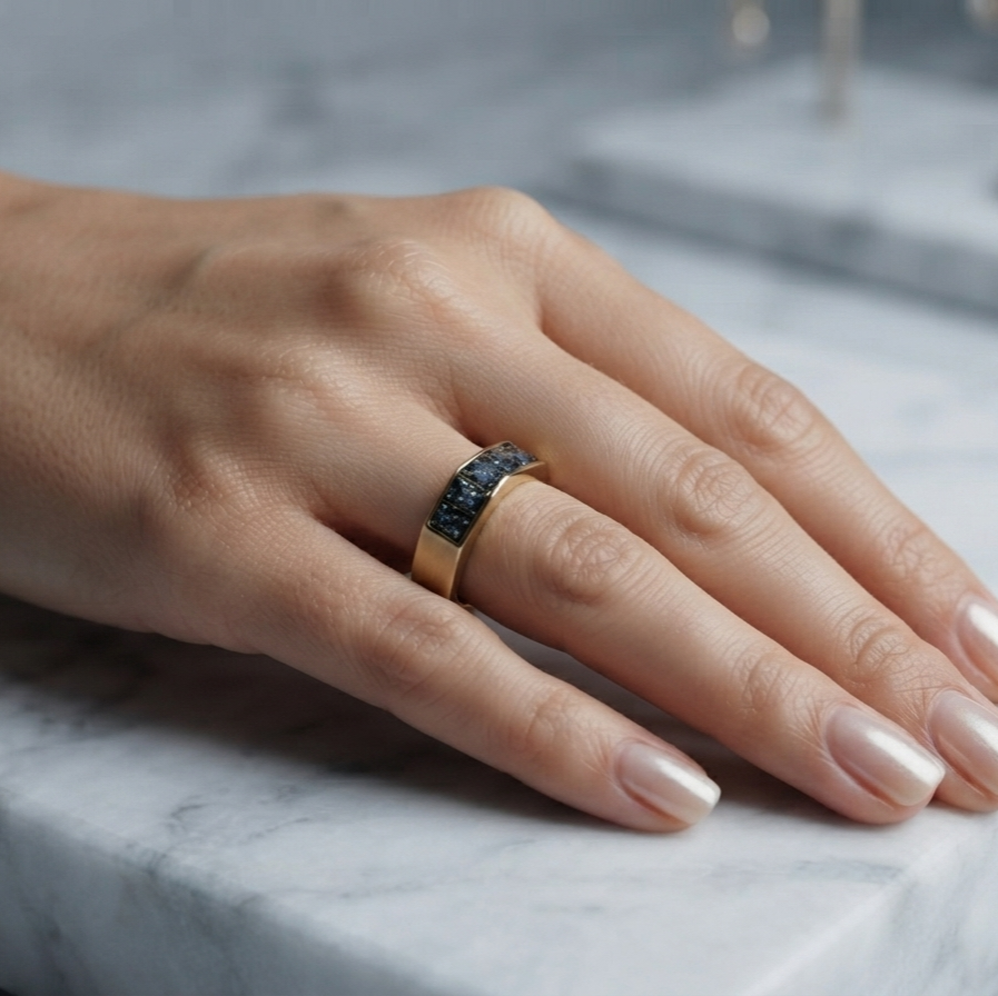 A close-up of a hand wearing a wide gold band ring with a dark textured inlay, resting on a white marble surface.