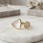 A gold ring with a triangular bezel-set clear stone and a round pavé-set clear stone, displayed on a white marble round tray.