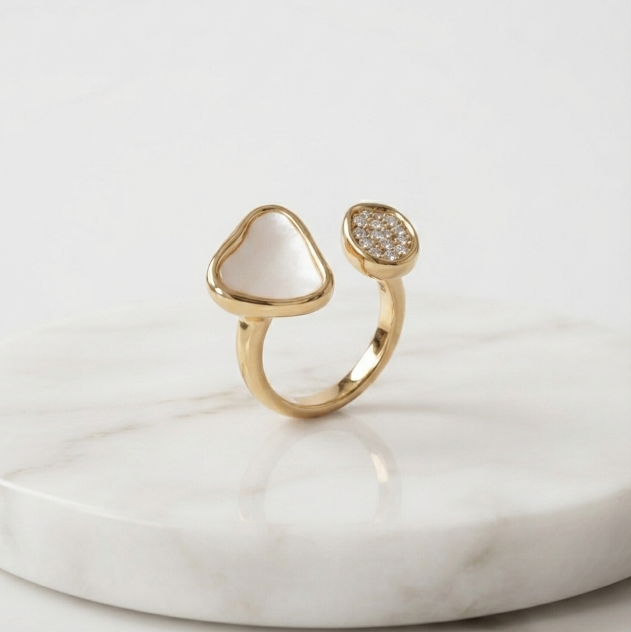 A gold open ring with one heart-shaped bezel containing a white inlay and the other featuring a pavé diamond setting, displayed on a white marble slab.