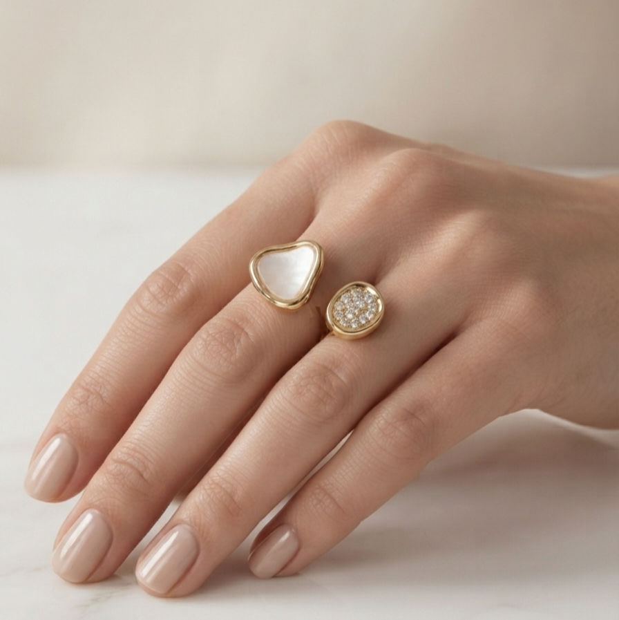 A close-up of a hand wearing two gold rings: one with a smooth white heart-shaped stone and the other with a textured white stone cluster, set against a soft beige background.