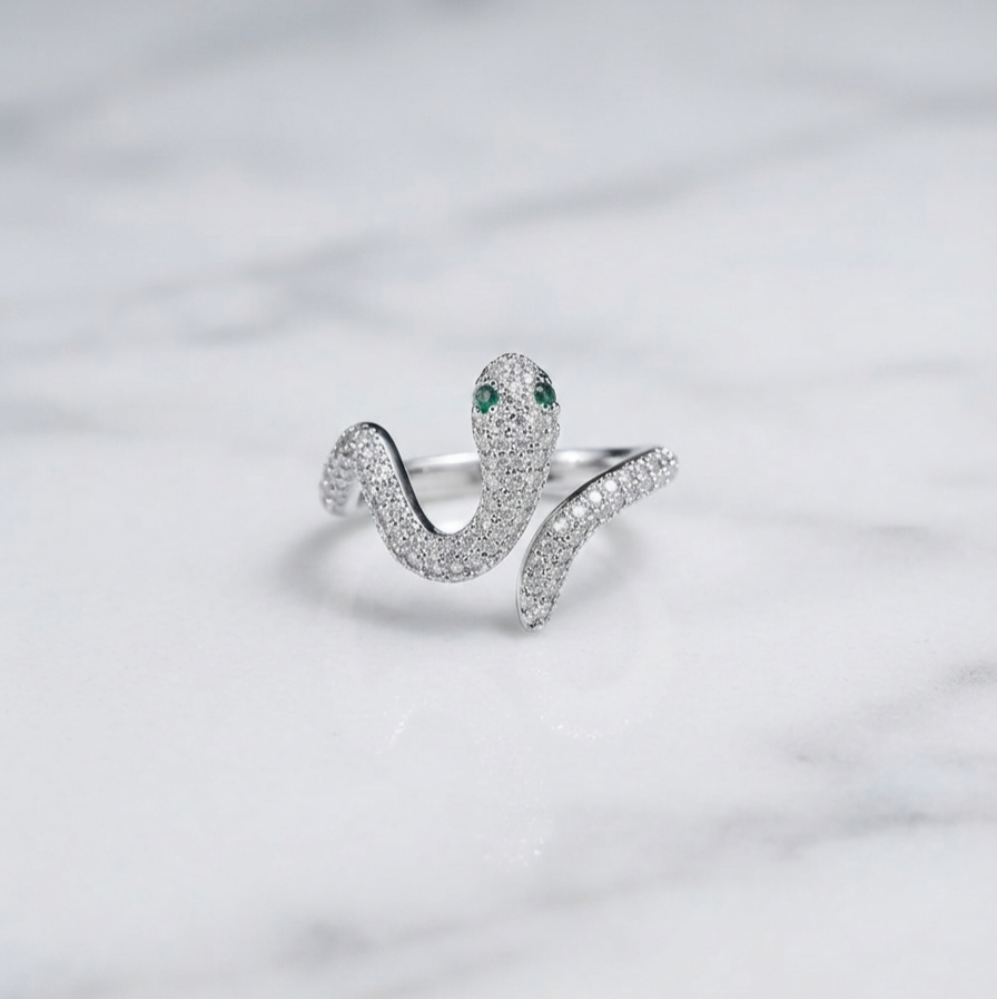 A silver snake ring paved with small clear stones, featuring green gemstone eyes, resting on a white marble surface.