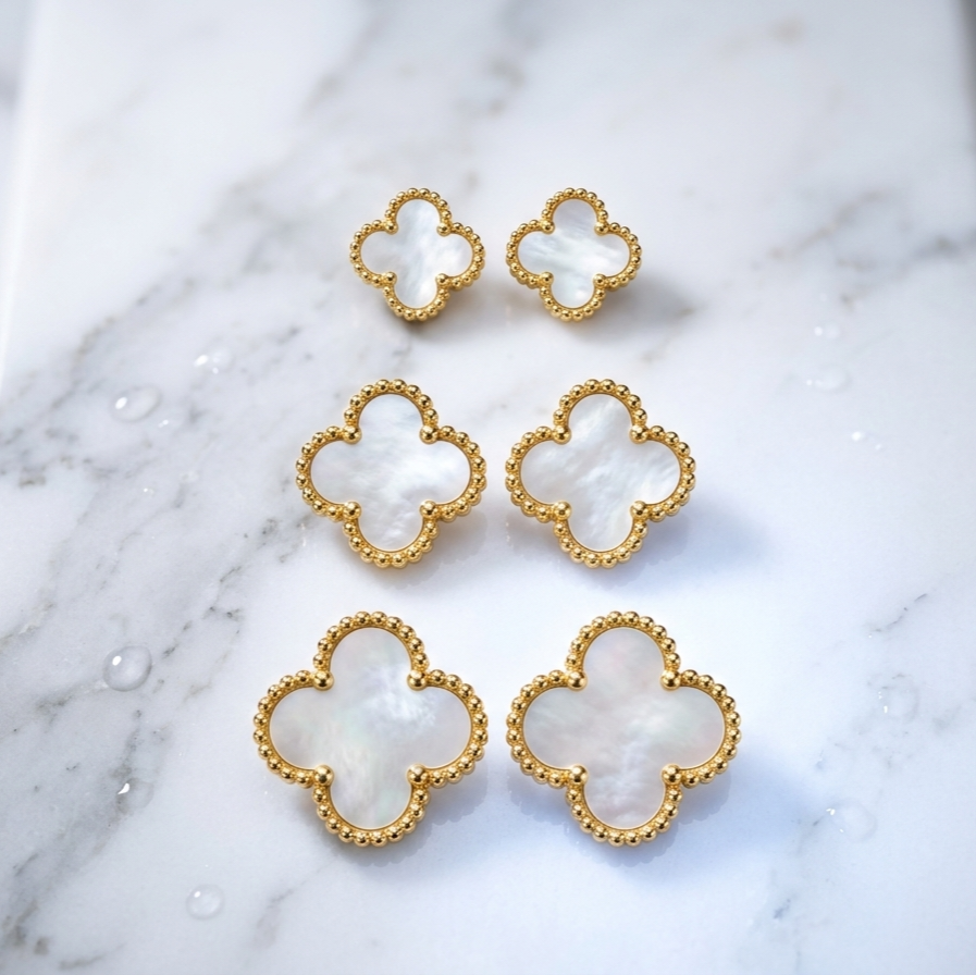 Six gold-tone four-leaf clover stud earrings with white mother-of-pearl inlays, arranged on a white marble surface.