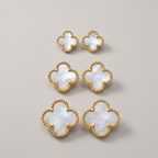 Six gold-tone four-leaf clover stud earrings with iridescent white mother-of-pearl centers, arranged on a neutral light beige background.
