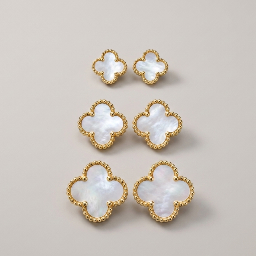 Six gold-tone four-leaf clover stud earrings with iridescent white mother-of-pearl centers, arranged on a neutral light beige background.