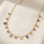 A gold chain necklace with multiple colorful gemstone charms, displayed on a textured cream linen fabric background.