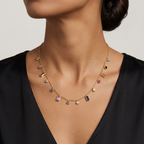 A close-up of a model wearing a delicate gold necklace with multiple small colorful gemstone charms and letter-shaped accents, paired with a black top against a light gray background.