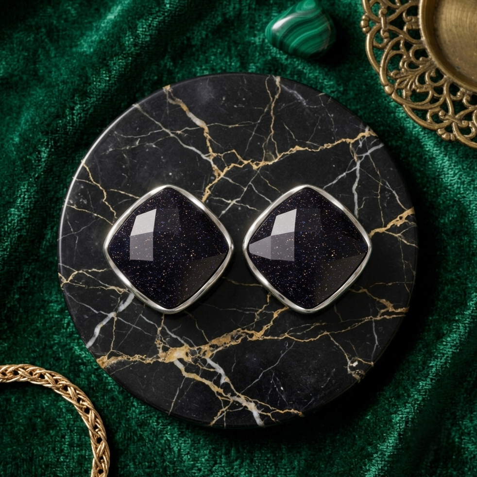 A pair of diamond-shaped stud earrings with dark blue glittery gemstones and silver settings, displayed on a round black marble coaster over a green velvet fabric background.