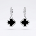 A pair of silver-toned hoop earrings with dangling clover-shaped charms. Each charm features a black inlay in the center and is surrounded by a halo of small clear stones, set against a clean white background.