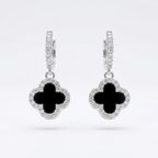 A pair of silver-toned hoop earrings with dangling clover-shaped charms. Each charm features a black inlay in the center and is surrounded by a halo of small clear stones, set against a clean white background.