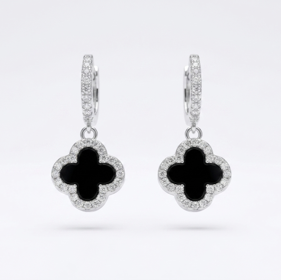 A pair of silver-toned hoop earrings with dangling clover-shaped charms. Each charm features a black inlay in the center and is surrounded by a halo of small clear stones, set against a clean white background.