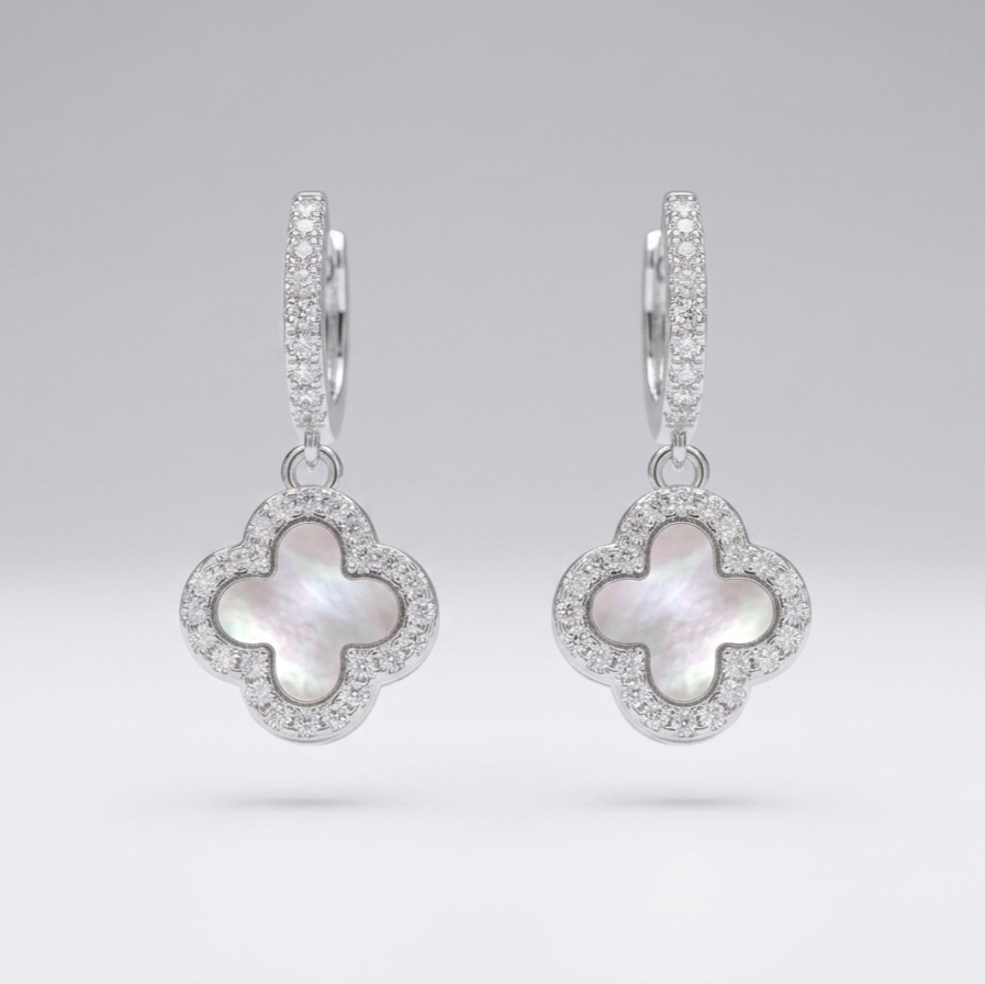 A pair of silver-toned dangle earrings featuring a clover motif with a white mother-of-pearl center and a border of small clear pavé stones, shown on a neutral light gray background.