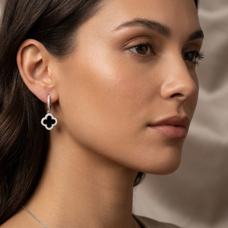A close-up side profile of a woman wearing a delicate silver-toned dangle earring with a black clover-shaped stone, set against a soft beige background.