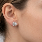 A close-up of a woman's ear wearing a round stud earring with a halo of small clear stones around a central round stone.