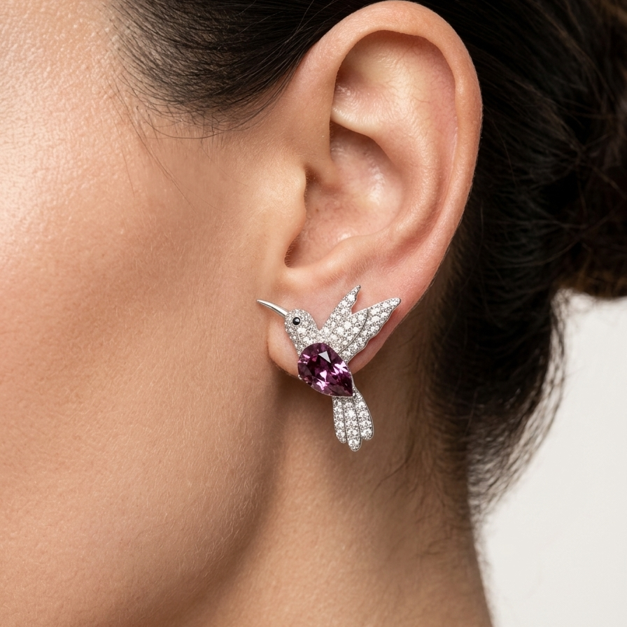 A close-up of a model's ear wearing a silver-toned bird stud earring with a purple heart-shaped gemstone and small clear stones.