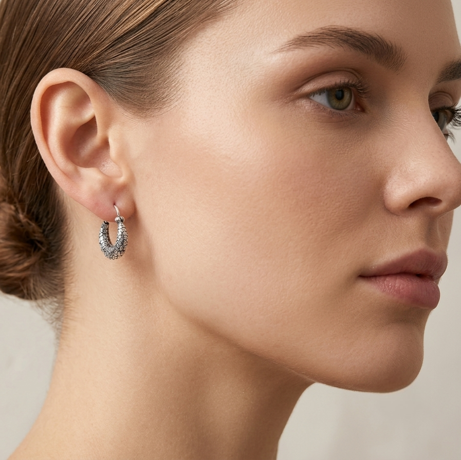 A close-up side profile of a woman wearing a small silver hoop earring with a textured, pavé-like surface that holds tiny clear stones. The background is a soft, neutral beige studio backdrop.