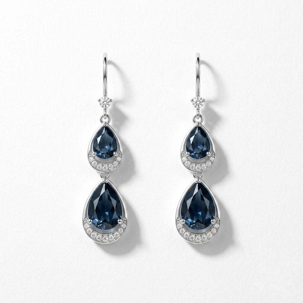 A pair of silver drop earrings featuring pear-shaped dark blue gemstones surrounded by small clear stones, shown on a clean white background.