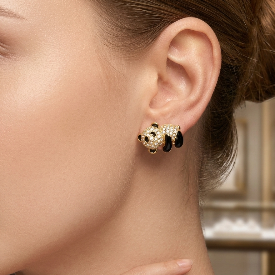 A close-up of a model's ear wearing a gold panda stud earring adorned with clear crystals and black enamel, with a softly blurred jewelry store background.