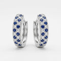 A pair of silver huggie hoop earrings adorned with rows of round-cut blue sapphires and clear pavé diamonds, set against a clean white background.