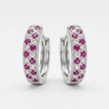 A pair of silver huggie hoop earrings decorated with pavé-set pink gemstones and small clear stones, shown against a light gray background.