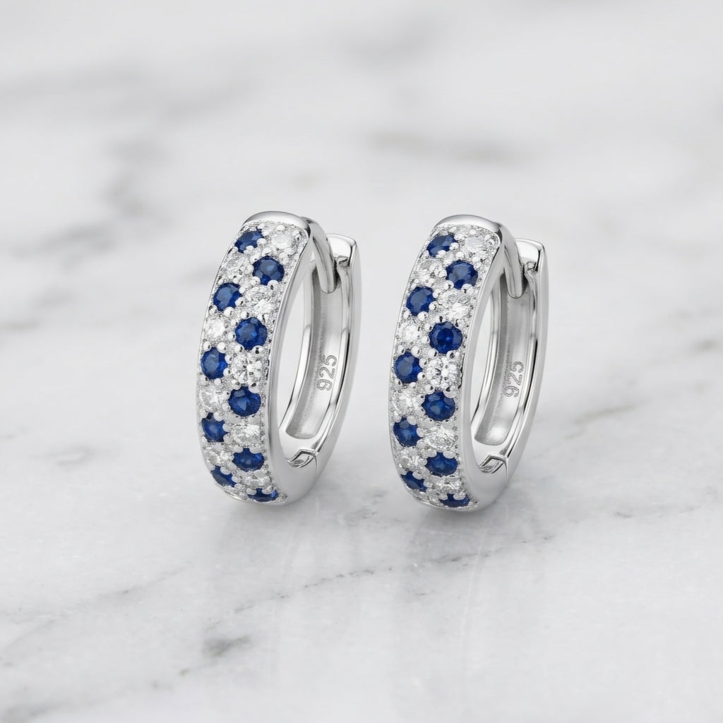 A pair of silver huggie hoop earrings set with blue sapphires and small clear pavé stones, displayed on a white marble surface. The inside of the earrings shows a '925' sterling silver hallmark.