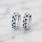 A pair of silver huggie hoop earrings set with blue sapphires and small clear pavé stones, displayed on a white marble surface. The inside of the earrings shows a '925' sterling silver hallmark.
