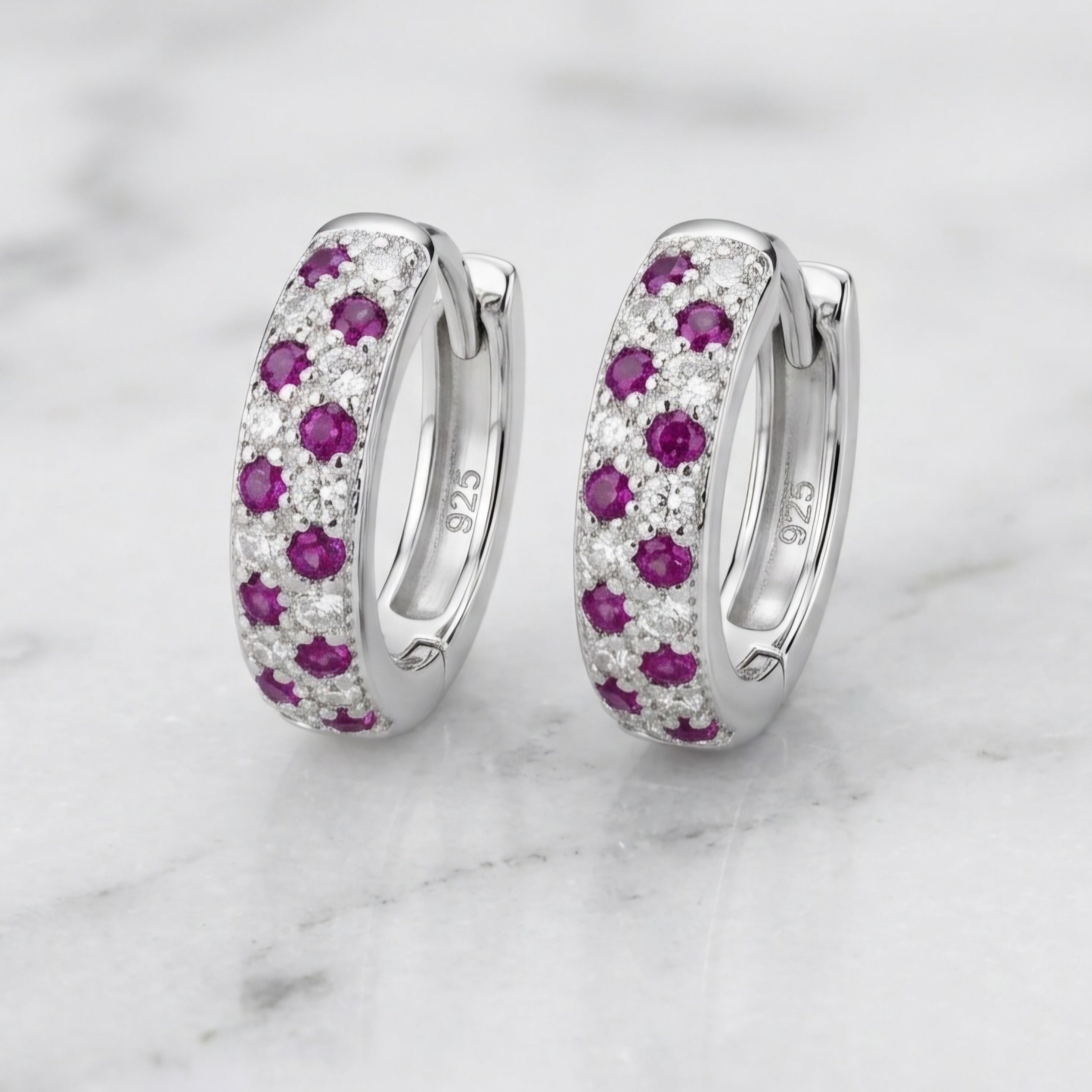 A pair of silver huggie hoop earrings with rows of round-cut pinkish-purple gemstones and clear pavé stones, displayed on a white marble surface.