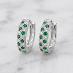 A pair of silver huggie hoop earrings decorated with rows of green emeralds and clear pavé stones, displayed on a white marble surface.