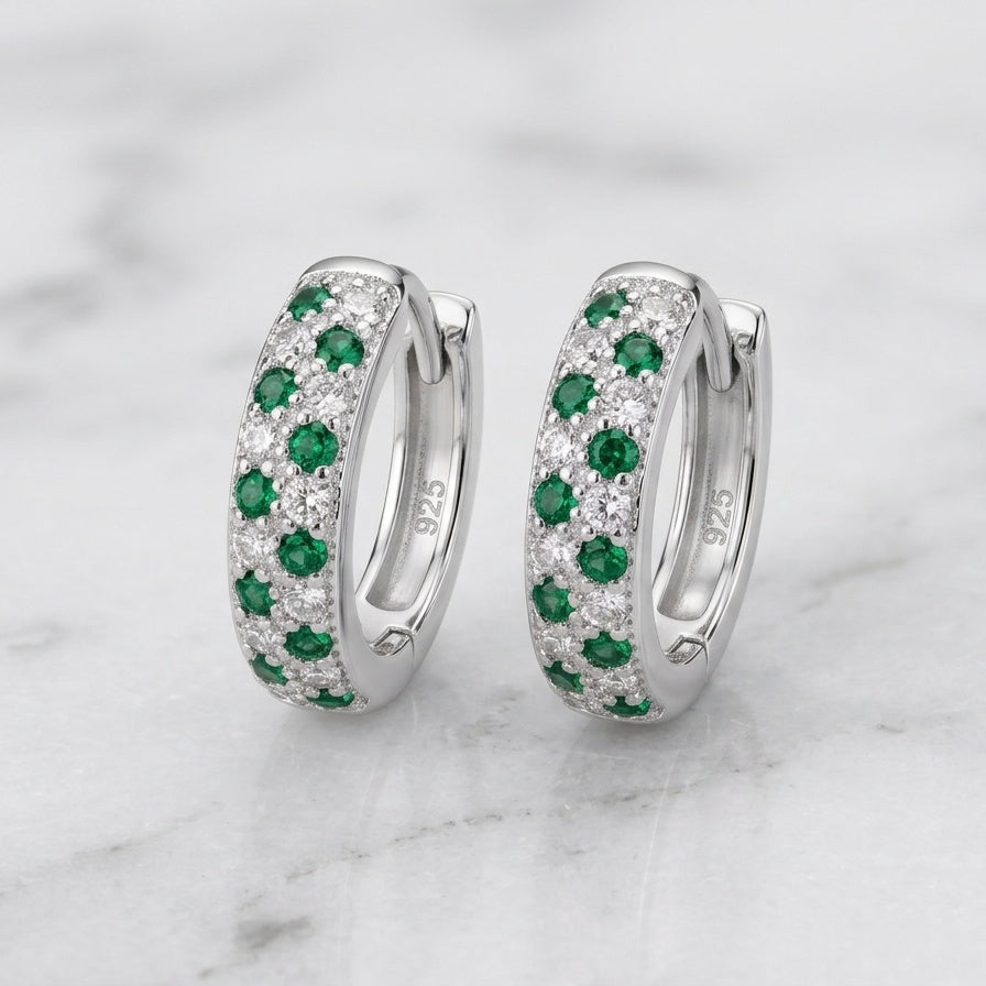 A pair of silver huggie hoop earrings decorated with rows of green emeralds and clear pavé stones, displayed on a white marble surface.