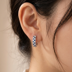 A close-up of a model's ear wearing a small silver huggie hoop earring with a blue and white patterned enamel face.