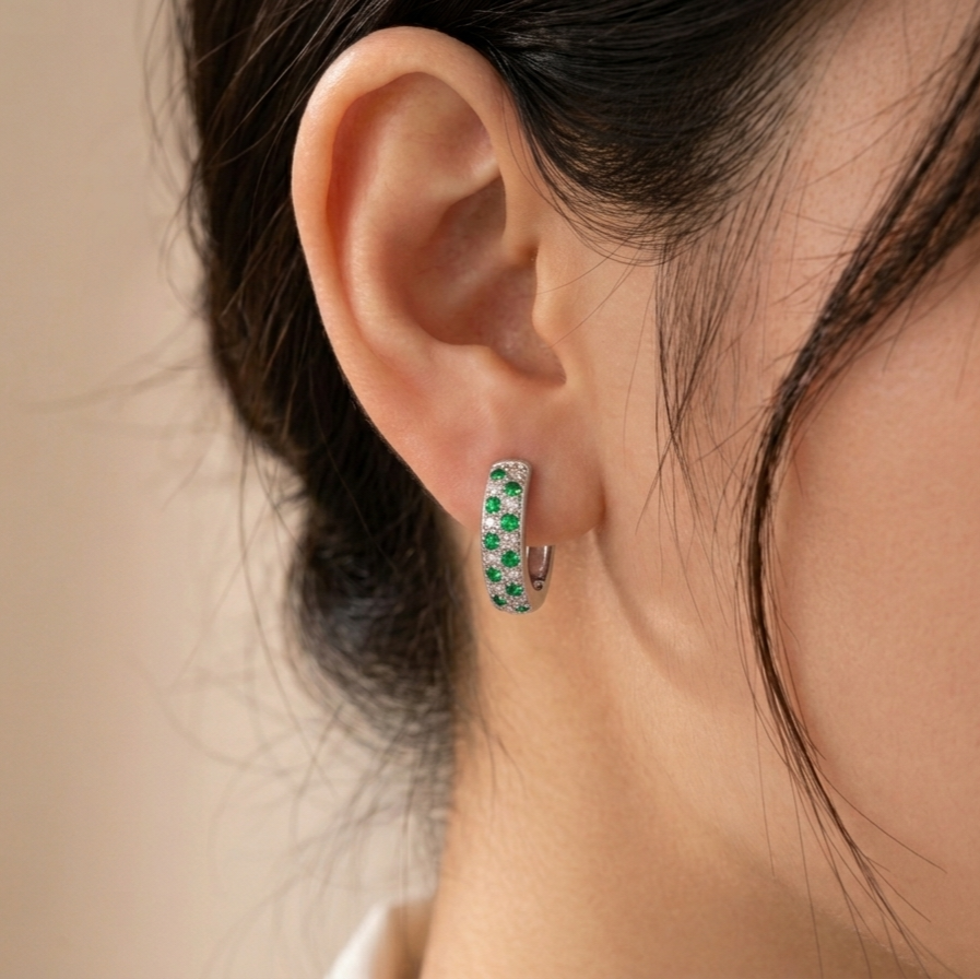 A close-up of a model's ear wearing a small silver huggie hoop earring set with green enamel and tiny white floral details.