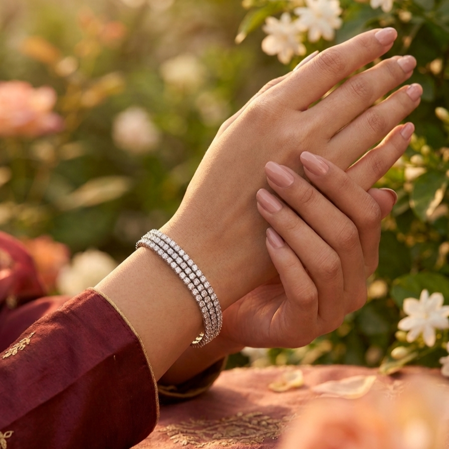 A close-up of a person's hands with manicured nails wearing a double-row diamond tennis bracelet, set against a soft-focus background of green foliage and white flowers.