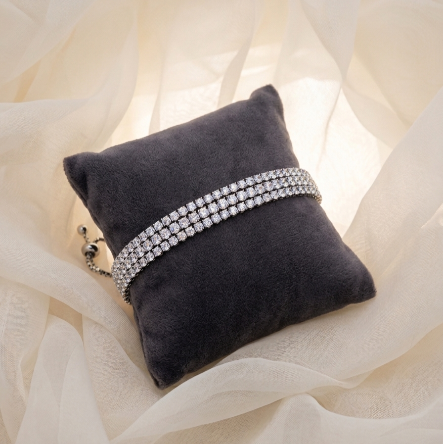 A double-row silver-toned bracelet with clear rhinestones displayed on a dark gray velvet pillow, set against a draped cream fabric background.
