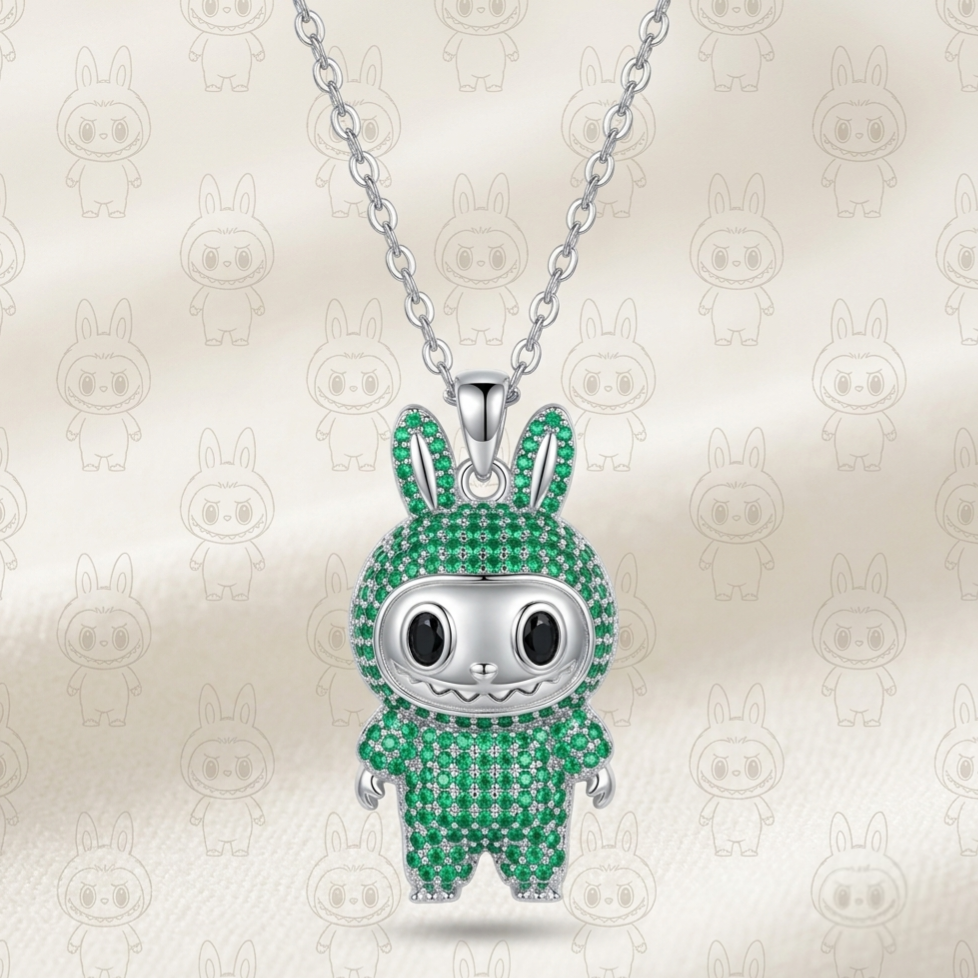 A silver bunny pendant necklace encrusted with green stones, hanging from a silver chain against a cream satin background with faint repeating bunny motifs.
