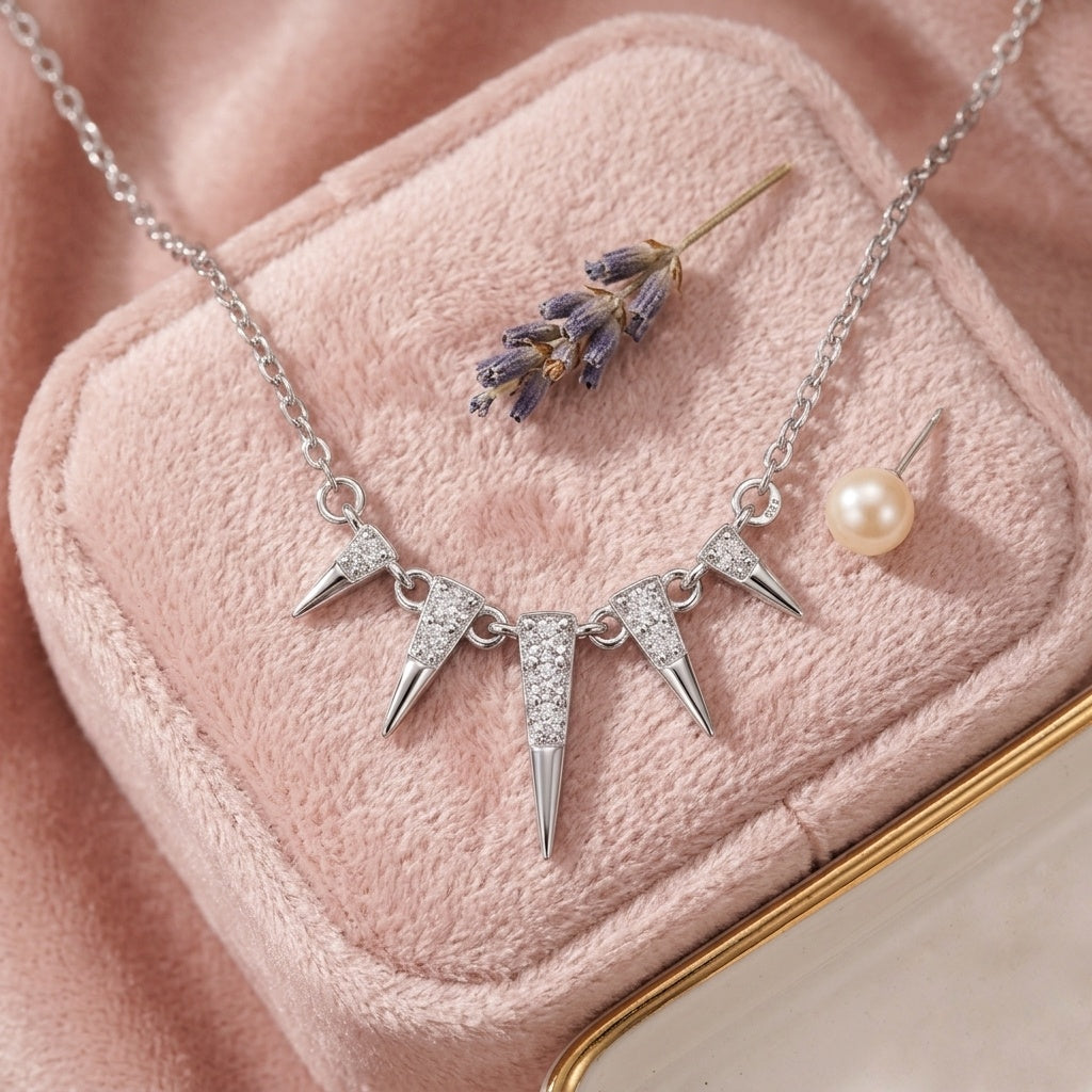 Silver-toned necklace with three spiked starburst pendants, displayed on a pink velvet jewelry box alongside a pearl stud earring and a small lavender sprig.