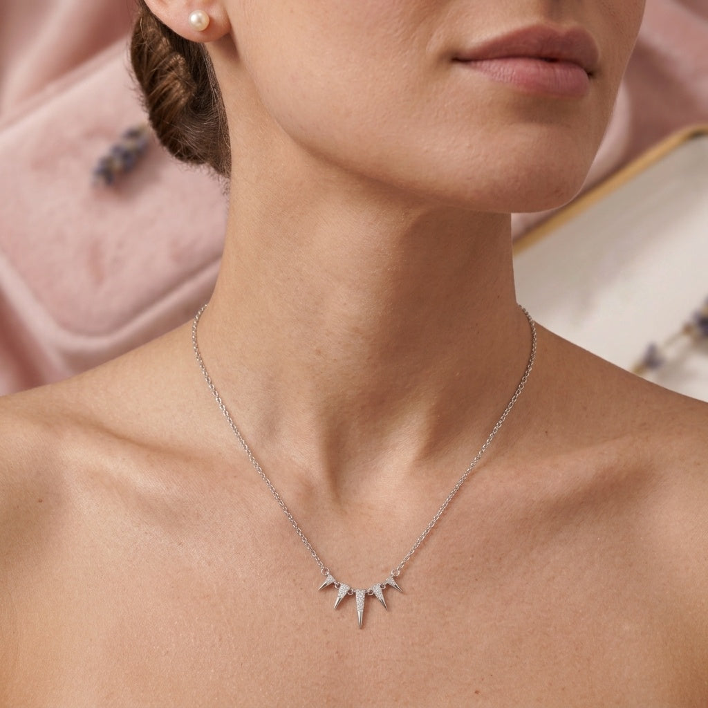 A close-up of a model wearing a delicate silver necklace with a multi-pointed starburst pendant, styled with a pearl earring against a soft pink background.