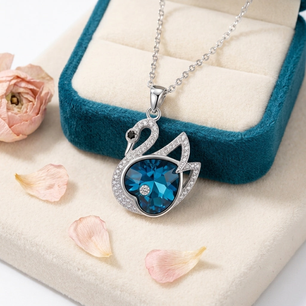 A silver-toned necklace with a swan-shaped pendant featuring a large faceted blue gemstone and small clear stones, displayed on a cream jewelry box with a teal velvet interior and scattered pink petals.