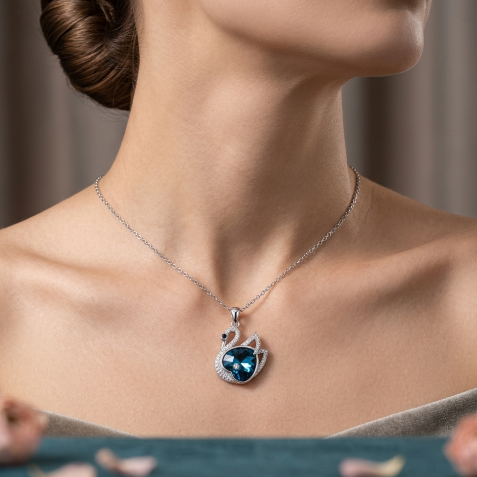 A close-up of a woman's neck and upper chest wearing a delicate silver-toned necklace with a swan pendant. The pendant features a dark blue heart-shaped center stone surrounded by small clear stones, with a curved neck detail.