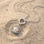 A silver-toned necklace with a shell pendant resting on sand. The pendant features a silver ring clasp, a silver scalloped shell outline, a white pearl in the center, and light blue enamel accents on the shell edges.
