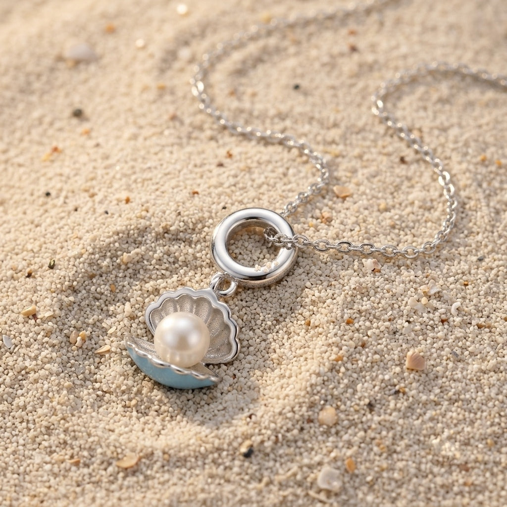 A silver-toned necklace with a shell pendant resting on sand. The pendant features a silver ring clasp, a silver scalloped shell outline, a white pearl in the center, and light blue enamel accents on the shell edges.