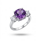 A silver three-stone ring featuring a large oval-cut purple amethyst center stone and two round clear accent stones on a white background.