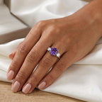 A close-up of a hand wearing a silver ring with a central round purple gemstone and small clear accent stones, resting on white satin fabric over a light wood surface.