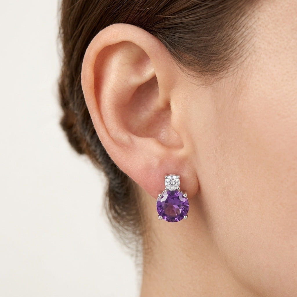 A close-up of a model's ear wearing a single stud earring with a round purple amethyst gemstone and a small square cluster of clear stones above it in a silver setting.