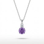 A silver pendant necklace featuring a round-cut purple amethyst gemstone in a four-prong setting, accented by a small cluster of clear stones above, suspended from a delicate silver chain against a white background.