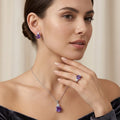 A woman models a matching jewelry set featuring a silver pendant necklace and stud earrings with deep purple teardrop-cut gemstones, set against a soft beige background.