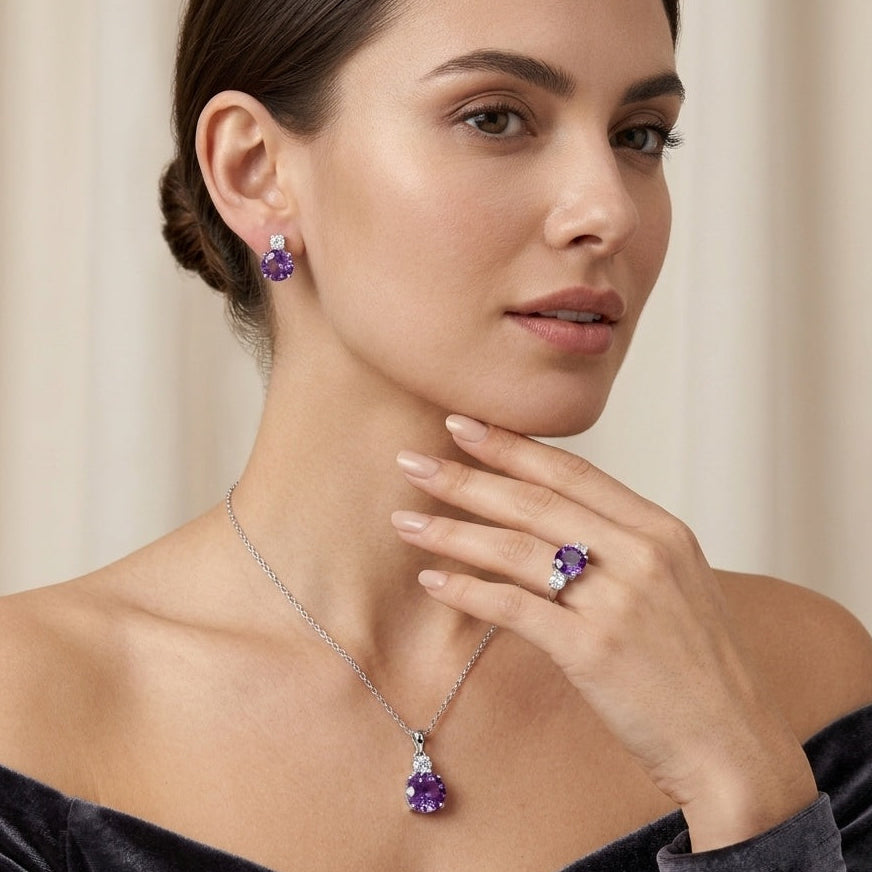 A woman models a matching jewelry set featuring a silver pendant necklace and stud earrings with deep purple teardrop-cut gemstones, set against a soft beige background.