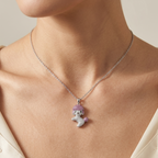 A close-up of a model wearing a delicate silver chain necklace with a small poodle pendant. The poodle is detailed with silver pavé stones and a soft lavender-purple crystal tuft for hair, set against a clean, neutral background.