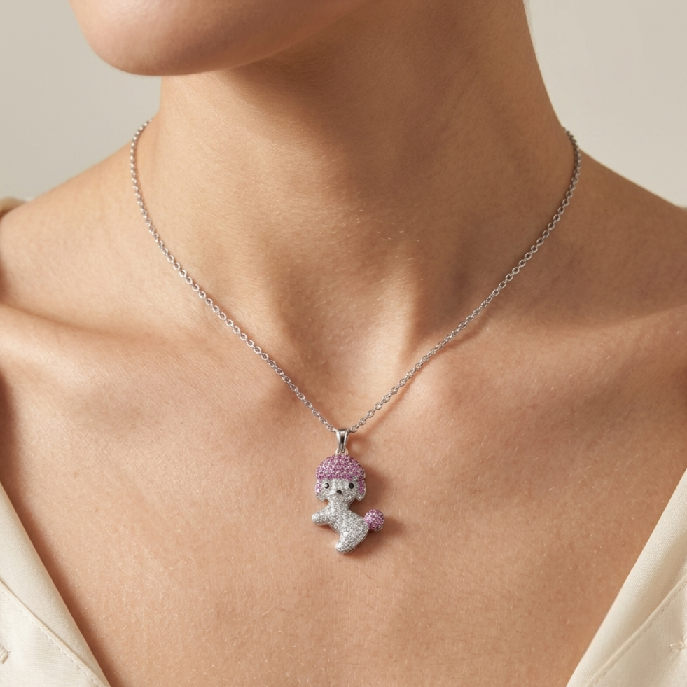 A close-up of a model wearing a delicate silver chain necklace with a small poodle pendant. The poodle is detailed with silver pavé stones and a soft lavender-purple crystal tuft for hair, set against a clean, neutral background.