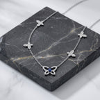 A silver-tone necklace with three small flower motifs and a larger butterfly pendant set with clear stones and a central blue gemstone, displayed on a dark gray marble block.