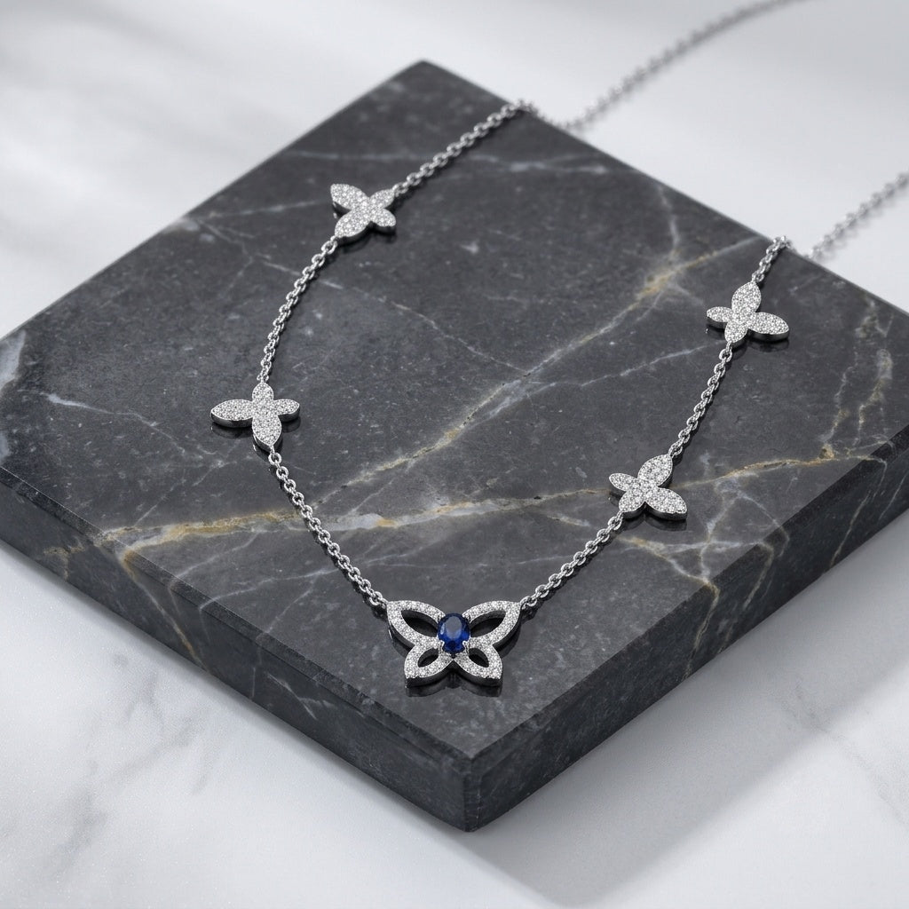 A silver-tone necklace with three small flower motifs and a larger butterfly pendant set with clear stones and a central blue gemstone, displayed on a dark gray marble block.